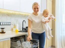 mother unloading baby bottles from a dishwasher while holding her smiling baby in a bright modern kitchen