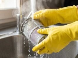 A woman using gloves to clean a cylindrical dishwasher filter under running hot water